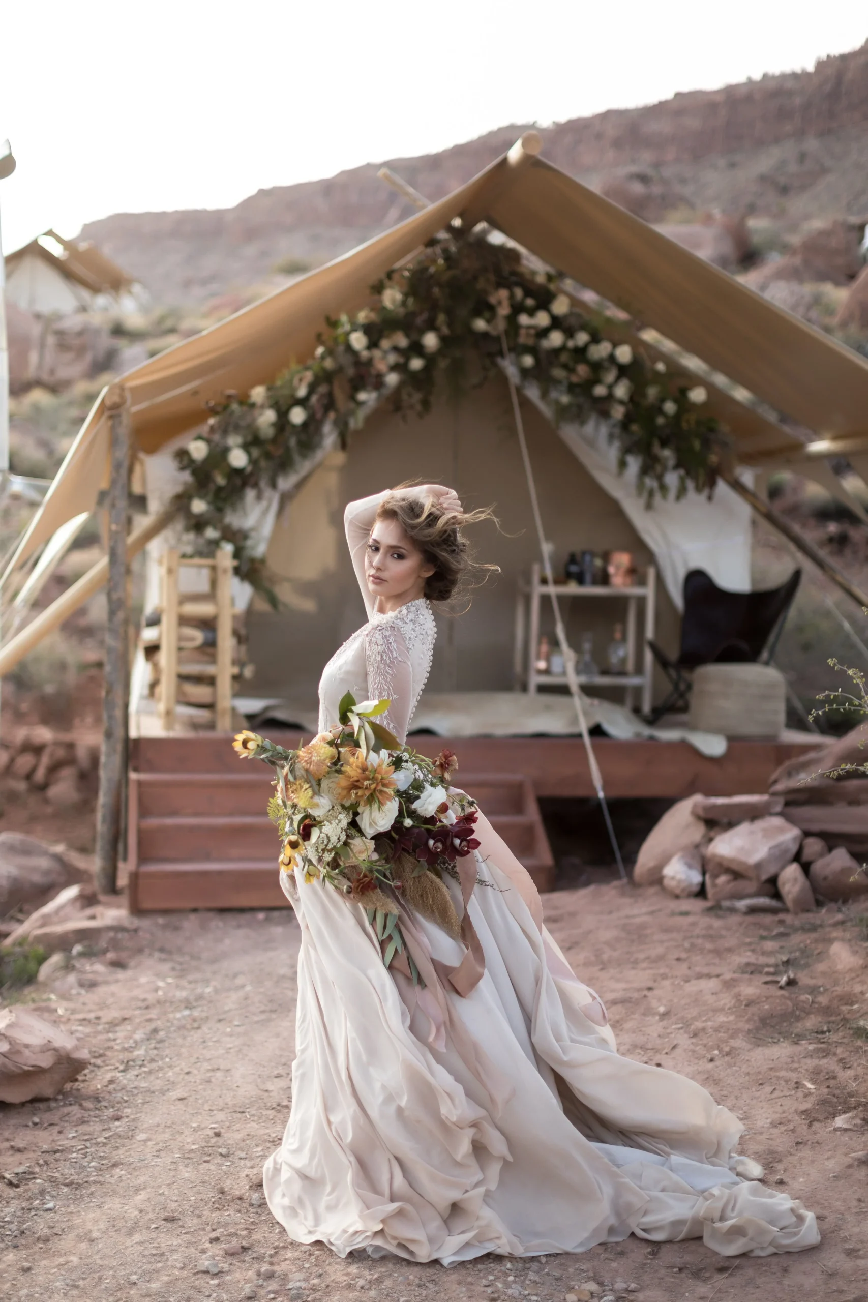 Bride posing in front of a tent with floral decorations, holding a large bouquet, outdoor wedding setting.