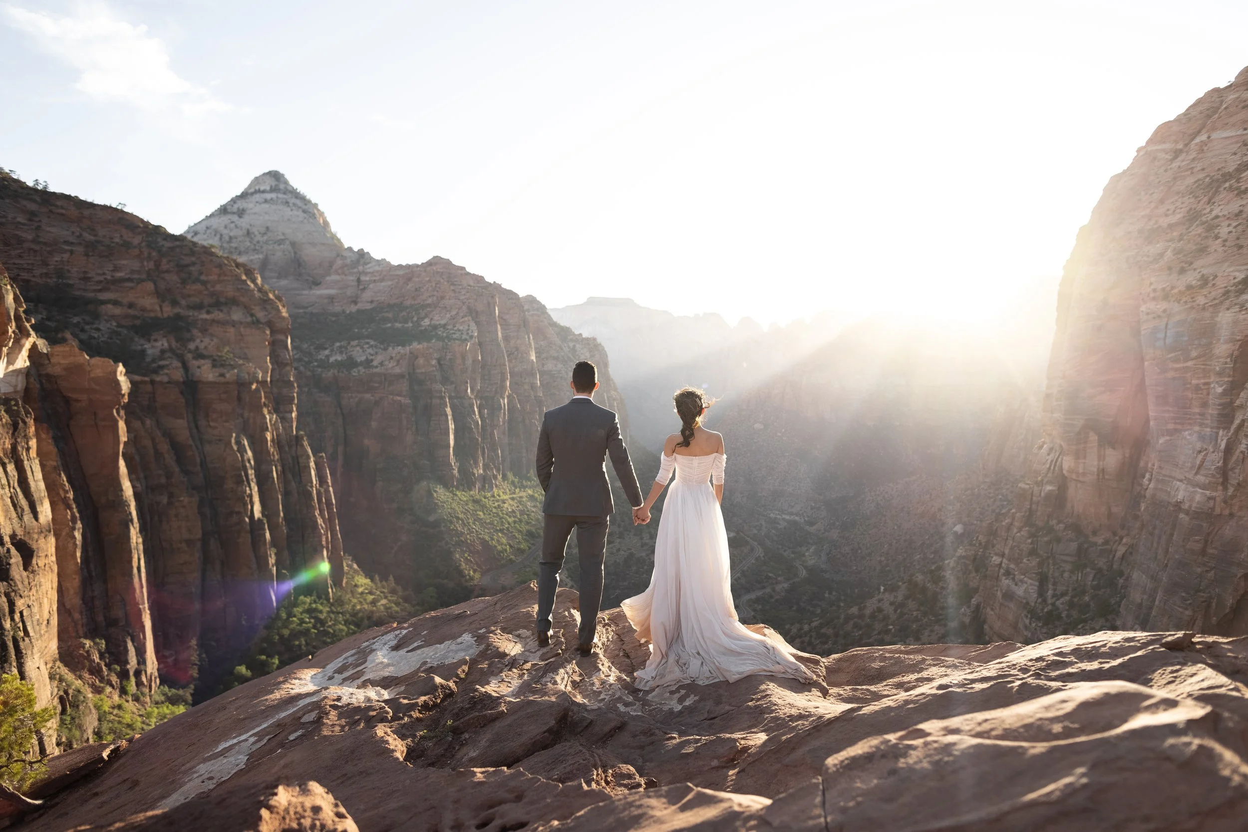 Bride and groom standing on a cliff overlooking a canyon with sunlight streaming in.
