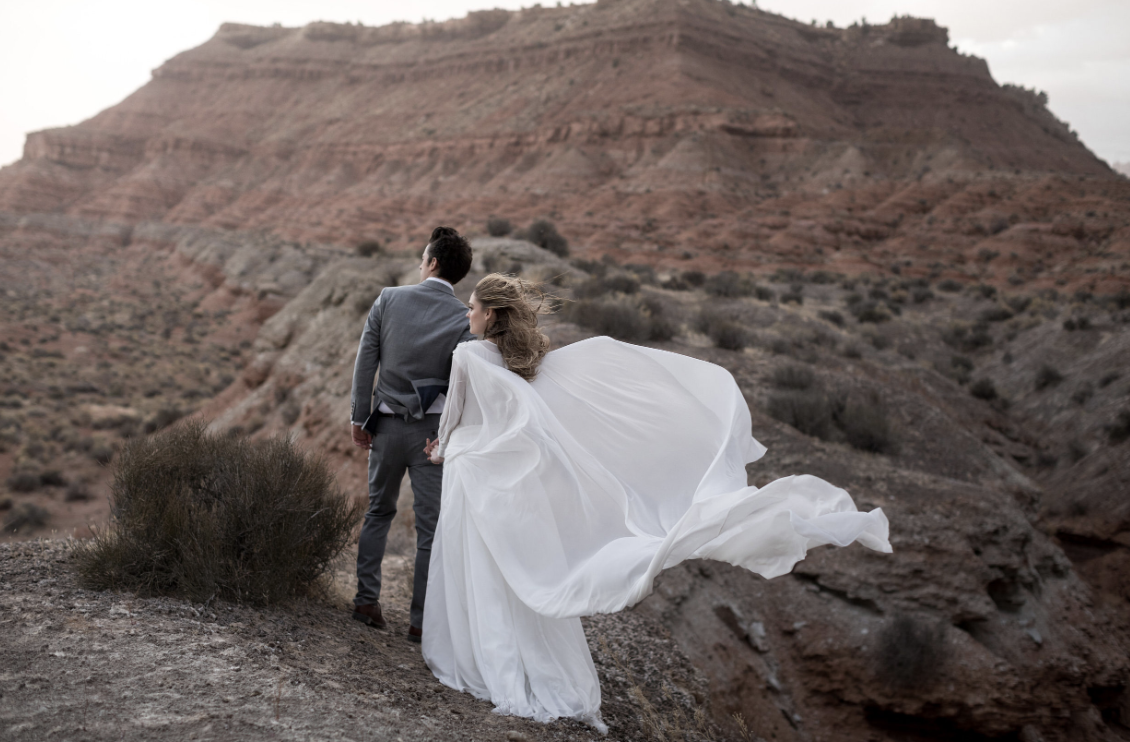 Bride and groom in desert landscape with flowing white dress and mountainous background.
