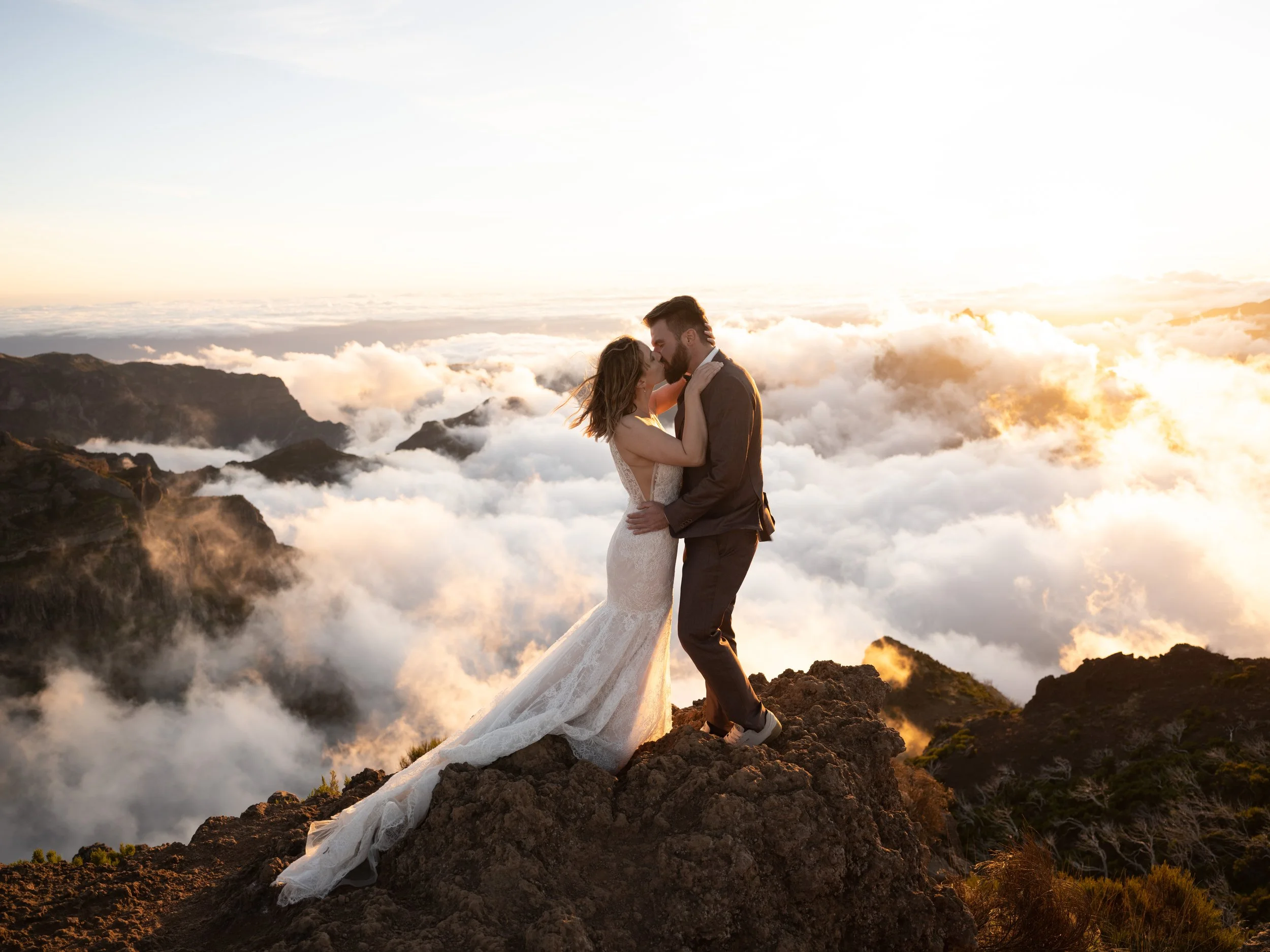 Couple in wedding attire kissing on a mountain peak above clouds at sunset.