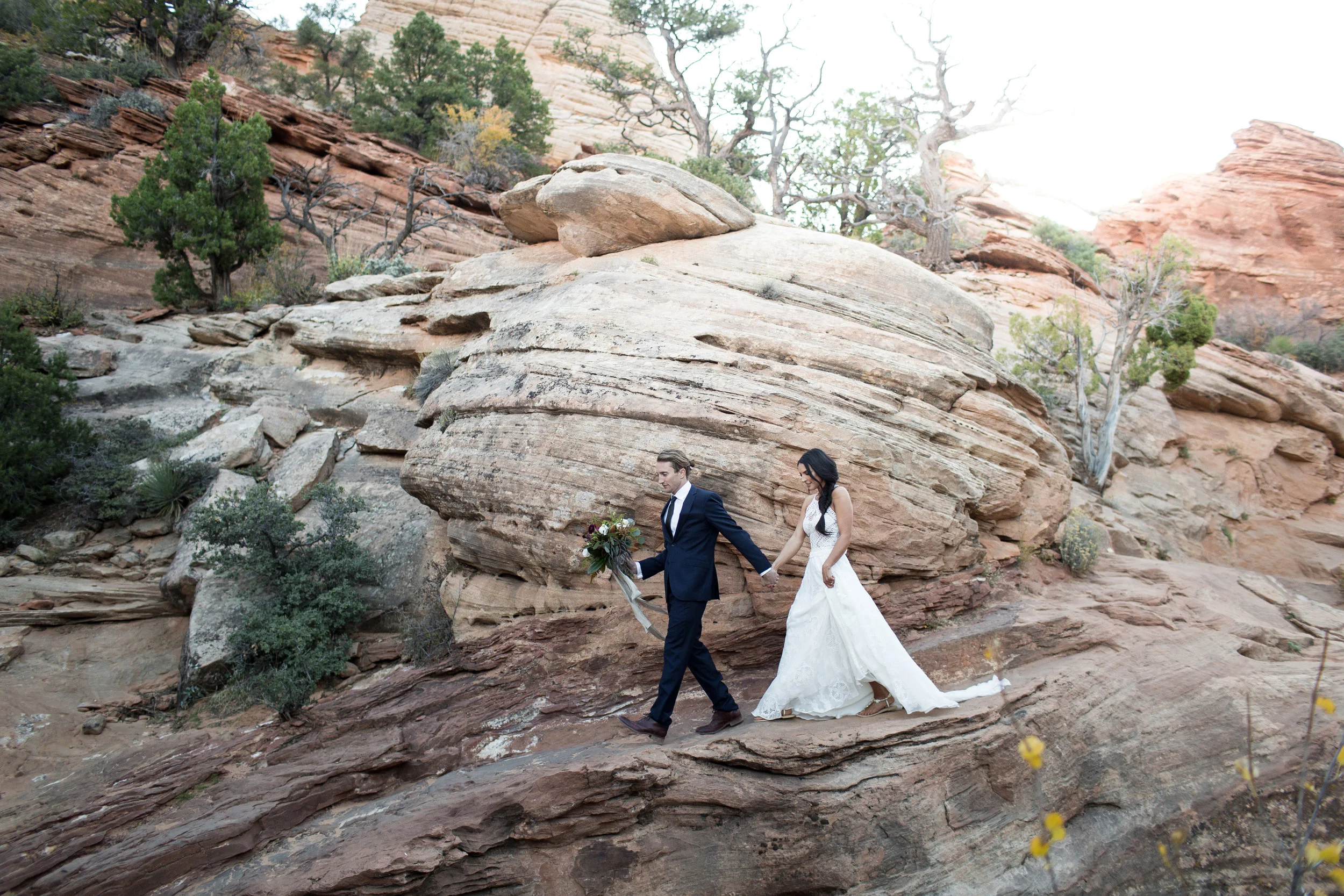 Bride and groom walking on rocky terrain in wedding attire, surrounded by desert landscape with trees and red rock formations.